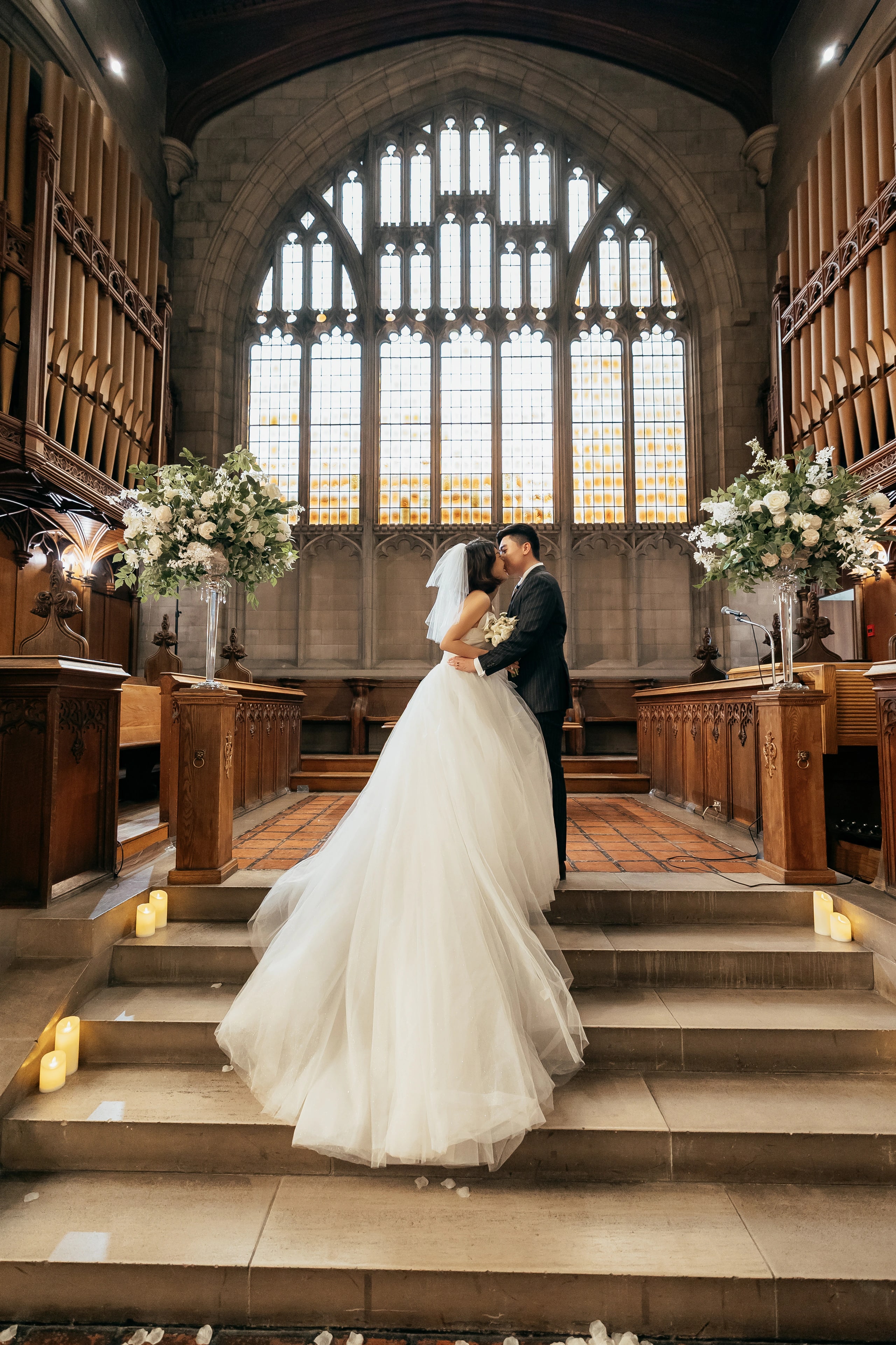 The couple at the altar steps beneath the great window, florals either side